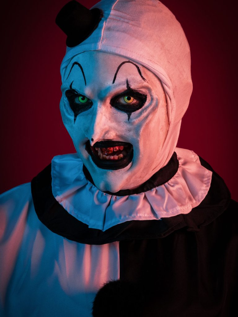 Portrait of Art the Clown from Terrifier with a black and white ruffled collar and small top hat against a red background.